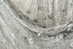 The image is a close-up of a fabric with a silver sequin pattern, featuring a mix of small and larger sequins, as well as some floral or leaf-like elements. The fabric appears to be made of a shiny, metallic material, giving it a sparkling and luxurious appearance.