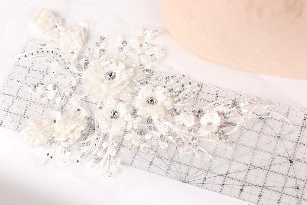a close-up view of a delicate, white lace flower with sparkling crystal elements. The flower is placed on a transparent ruler, which is lying on a white surface. The ruler appears to be used for measuring or cutting the lace, possibly for a craft or sewing project.