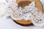 a close-up view of a delicate, intricate lace fabric with a floral pattern. The lace appears to be made of a soft, white material and is displayed on a wooden surface, possibly a piece of driftwood or a wooden board. The lace fabric is spread out, creating a visually appealing and elegant composition.