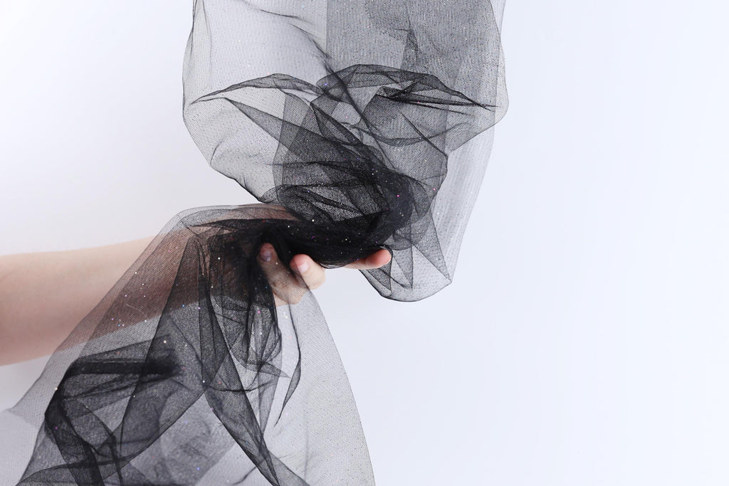 A hand is holding a black, sheer fabric with a sparkly texture, against a plain white background.