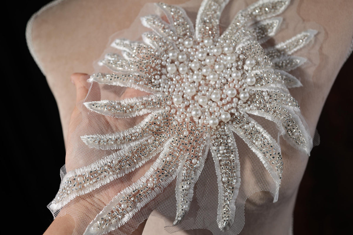 A hand is holding a decorative piece of fabric with a floral design made of white pearls and silver beads.