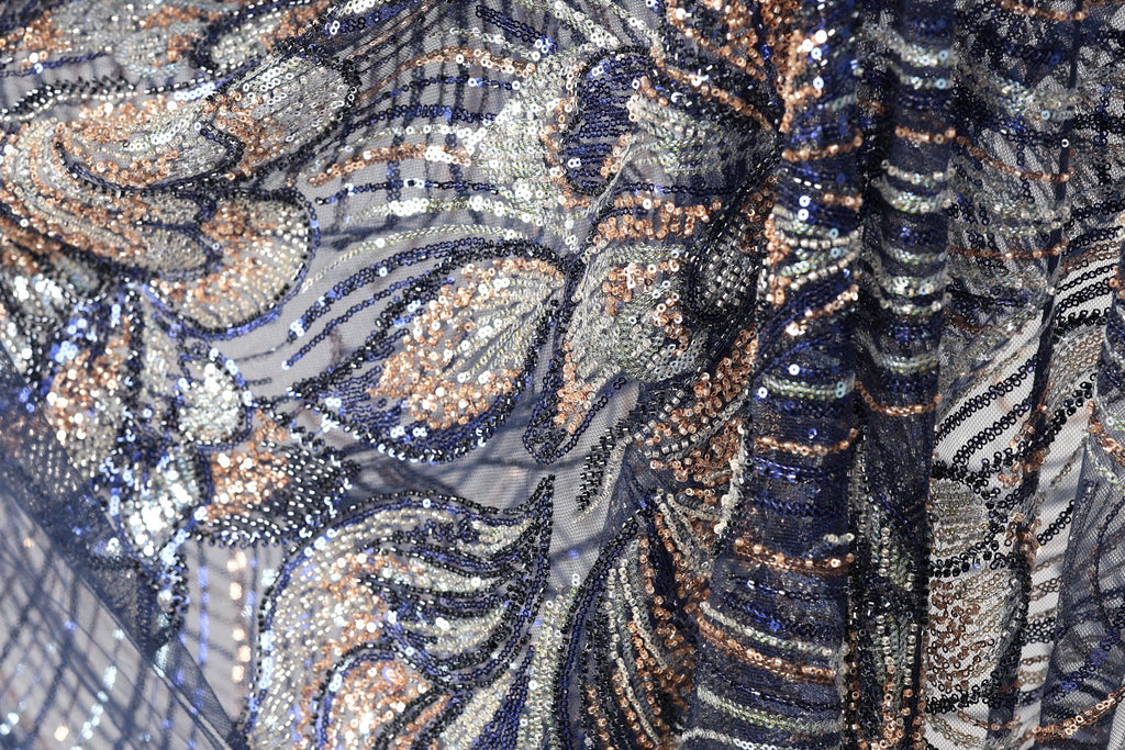 The image is a close-up of a fabric with a complex, intricate pattern of sequins and beads in shades of blue, gold, and silver. The pattern appears to be a floral or leaf-like design, with the sequins and beads arranged in a way that creates a visually striking and detailed appearance.