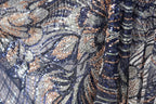 The image is a close-up of a fabric with a complex, intricate pattern of sequins and beads in shades of blue, gold, and silver. The pattern appears to be a floral or leaf-like design, with the sequins and beads arranged in a way that creates a visually striking and detailed appearance.