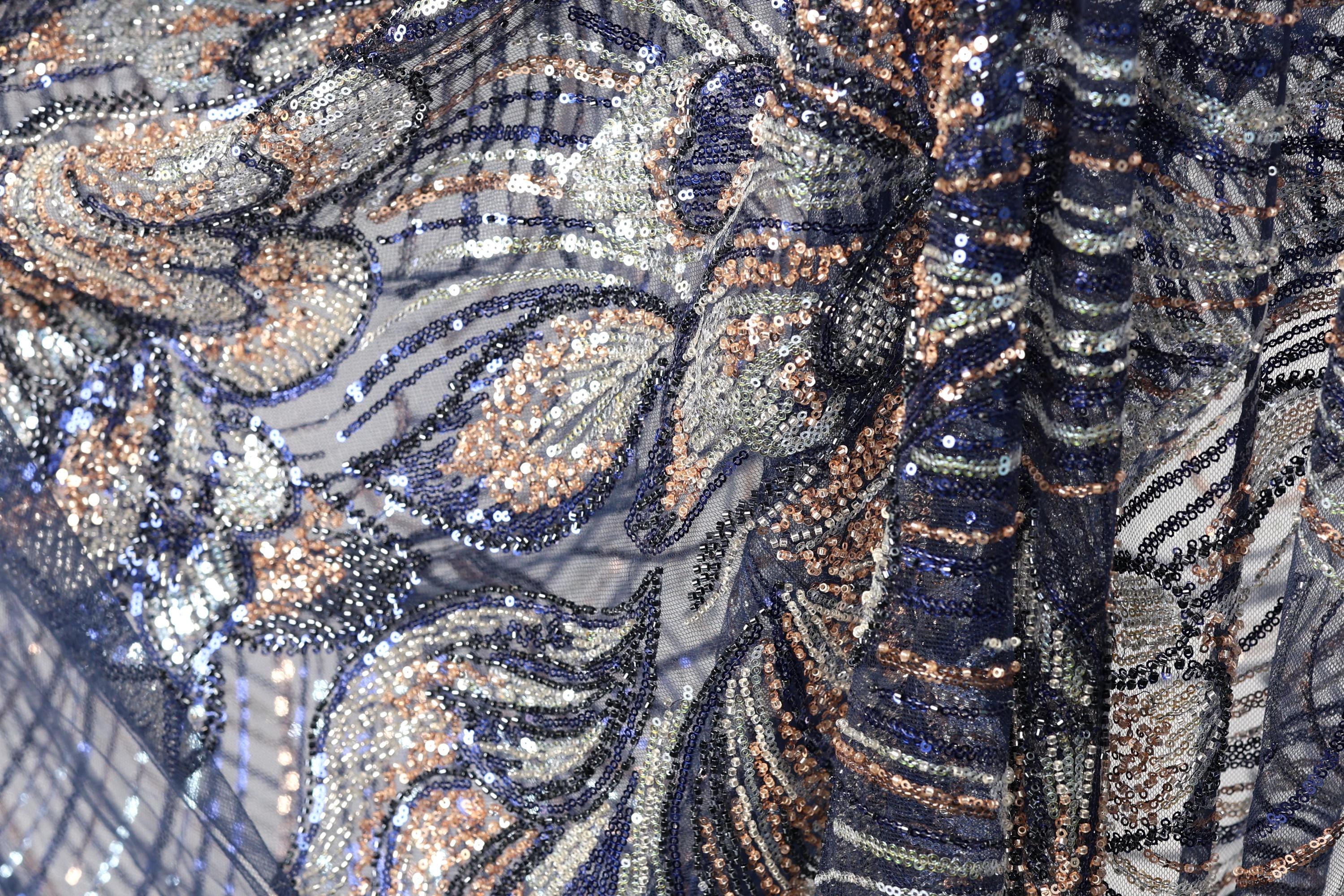 The image is a close-up of a fabric with a complex, intricate pattern of sequins and beads in shades of blue, gold, and silver. The pattern appears to be a floral or leaf-like design, with the sequins and beads arranged in a way that creates a visually striking and detailed appearance.