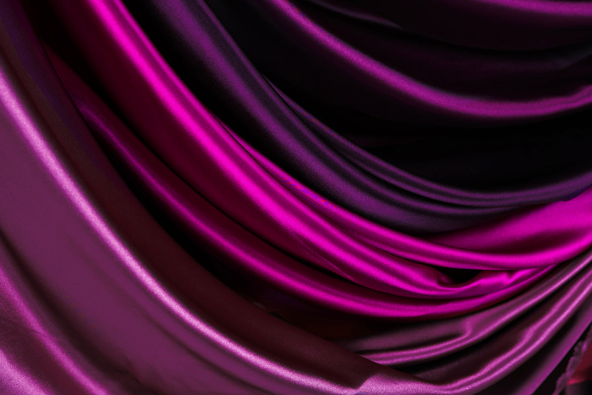 a close-up view of a fabric with a gradient of colors, transitioning from a deep purple to a lighter shade of purple. The fabric appears to be made of satin or a similar smooth, shiny material, and it is folded or draped in a way that creates a sense of depth and texture.