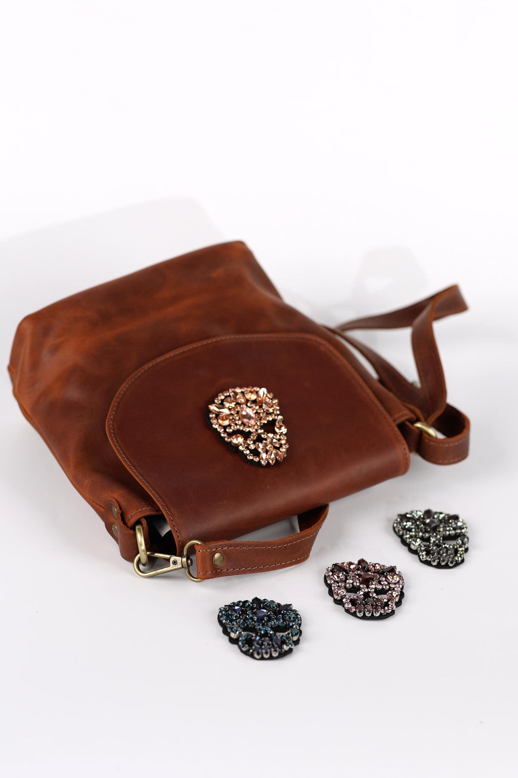 A brown leather handbag with a decorative skull-shaped embellishment on the front, accompanied by three small rhinestone-encrusted brooches.