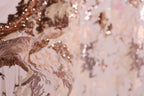 The image is a close-up of a fabric with a floral pattern, featuring a mix of pink and gold colors. The fabric appears to be made of lace or a similar delicate material, with intricate details and textures. The background is blurred, drawing focus to the fabric and its details.