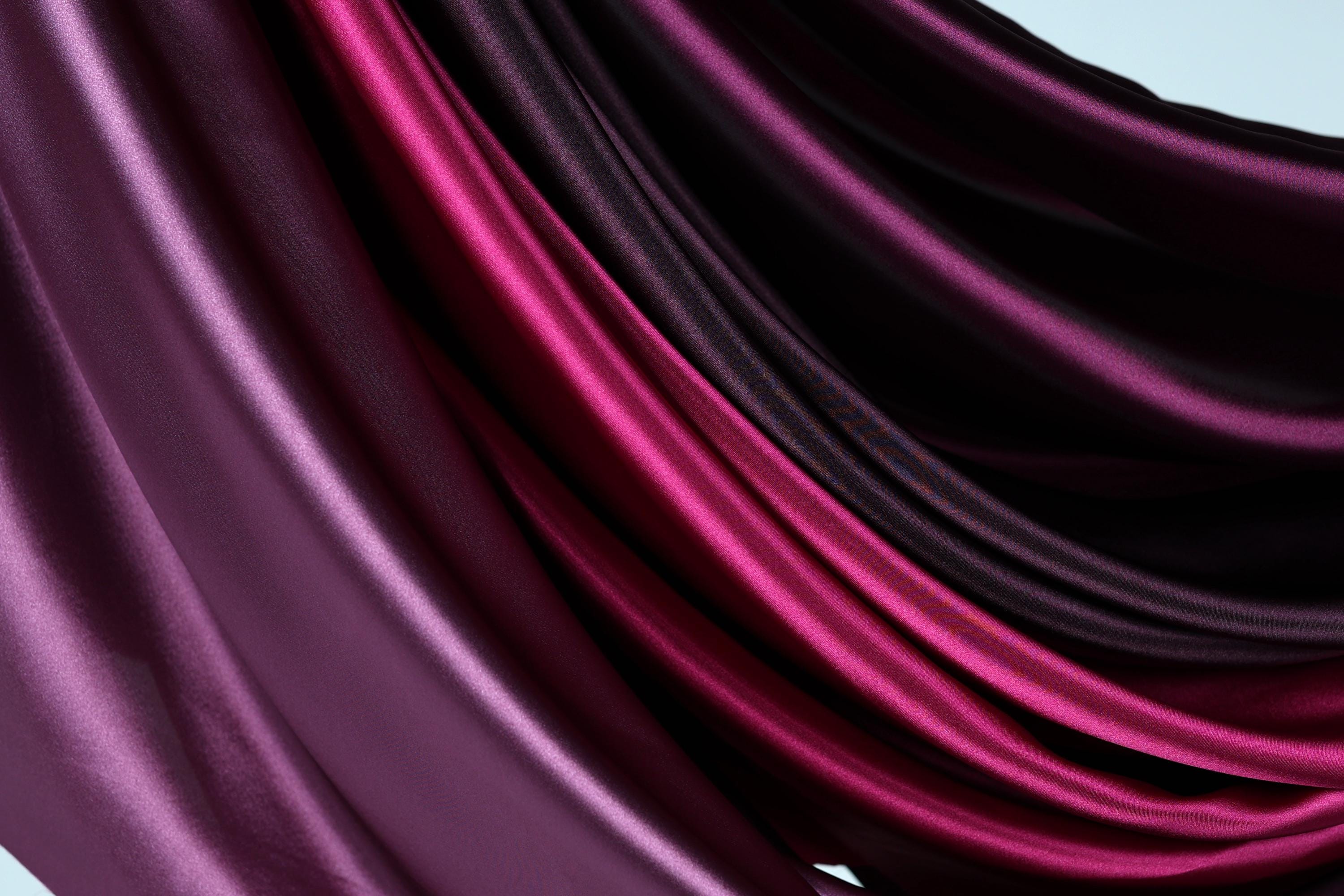 a close-up view of a fabric with a gradient of colors, transitioning from a deep purple to a lighter shade of purple. The fabric appears to be made of silk or satin, with a smooth and shiny texture. The colors are vibrant and distinct, creating a visually striking and elegant appearance.