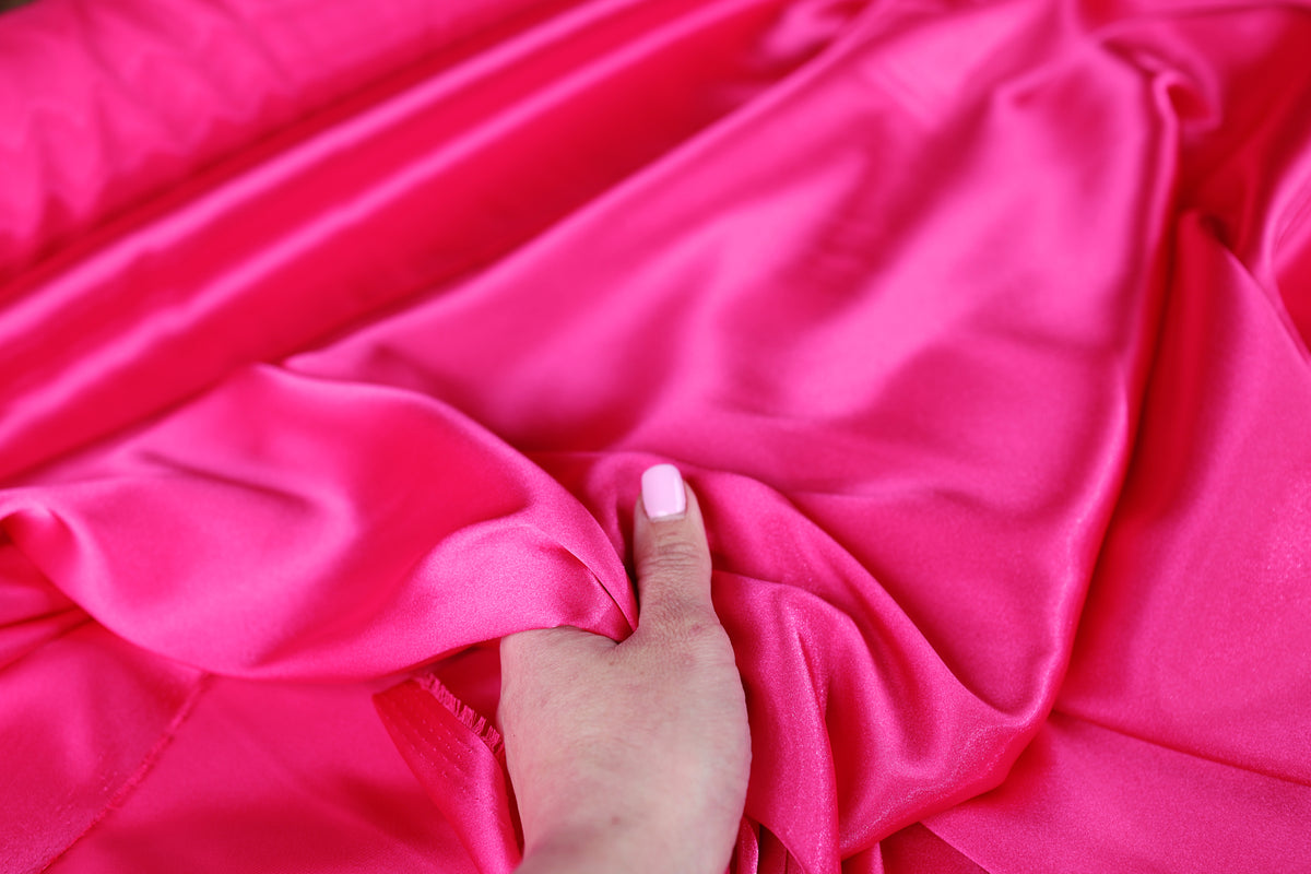 A hand is holding a piece of bright pink satin fabric, with the fabric's folds and creases visible.