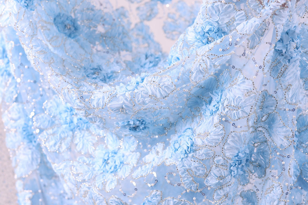 a close-up view of a delicate, light blue fabric with a floral pattern. The fabric appears to be made of a sheer material, with small white flowers or sequins scattered throughout the design. The overall appearance of the fabric is soft and delicate, with a sense of elegance and sophistication.