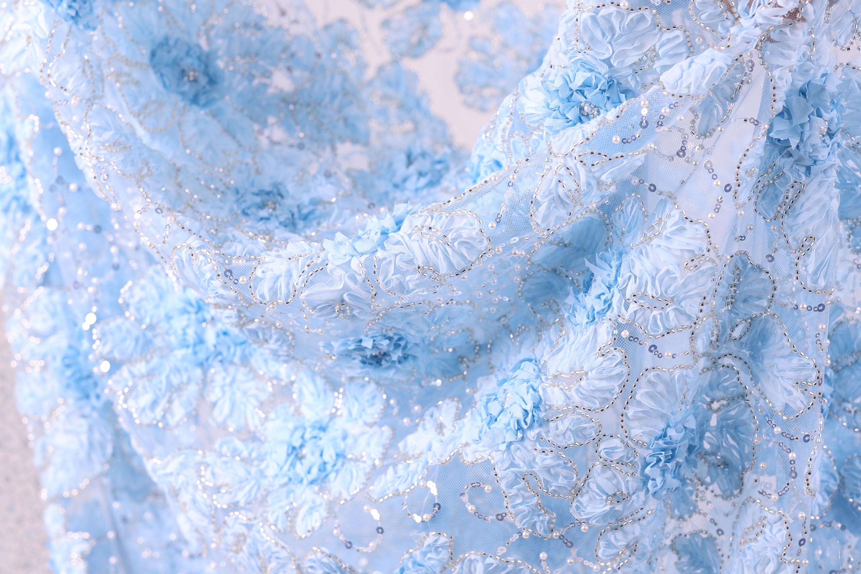 a close-up view of a delicate, light blue fabric with a floral pattern. The fabric appears to be made of a sheer material, with small white flowers or sequins scattered throughout the design. The overall appearance of the fabric is soft and delicate, with a sense of elegance and sophistication.