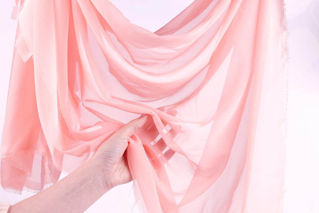 A hand is holding a pink fabric, possibly silk or chiffon, against a white background.