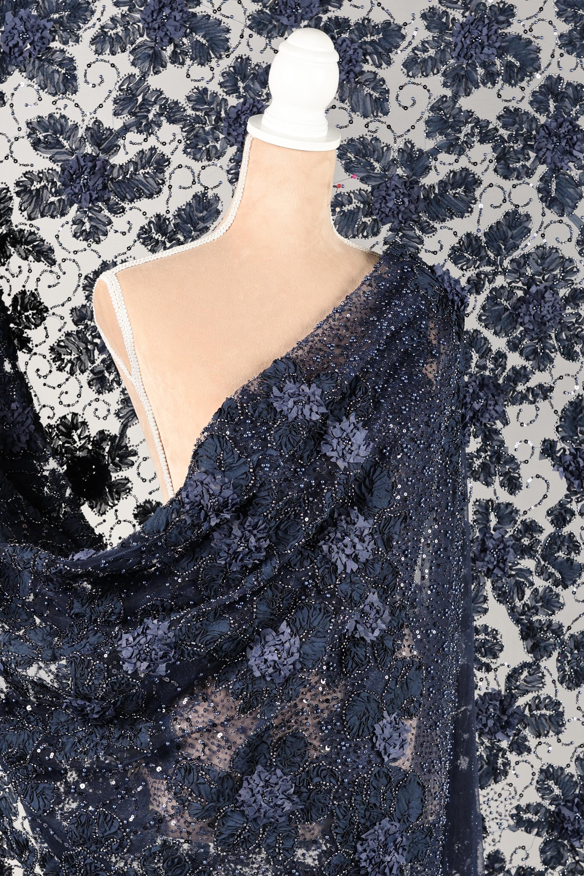 a close-up view of a piece of fabric with a floral pattern, featuring a lace-like texture. The fabric appears to be a dark blue color with white accents, and there is a white object, possibly a piece of clothing or accessory, partially visible in the frame.