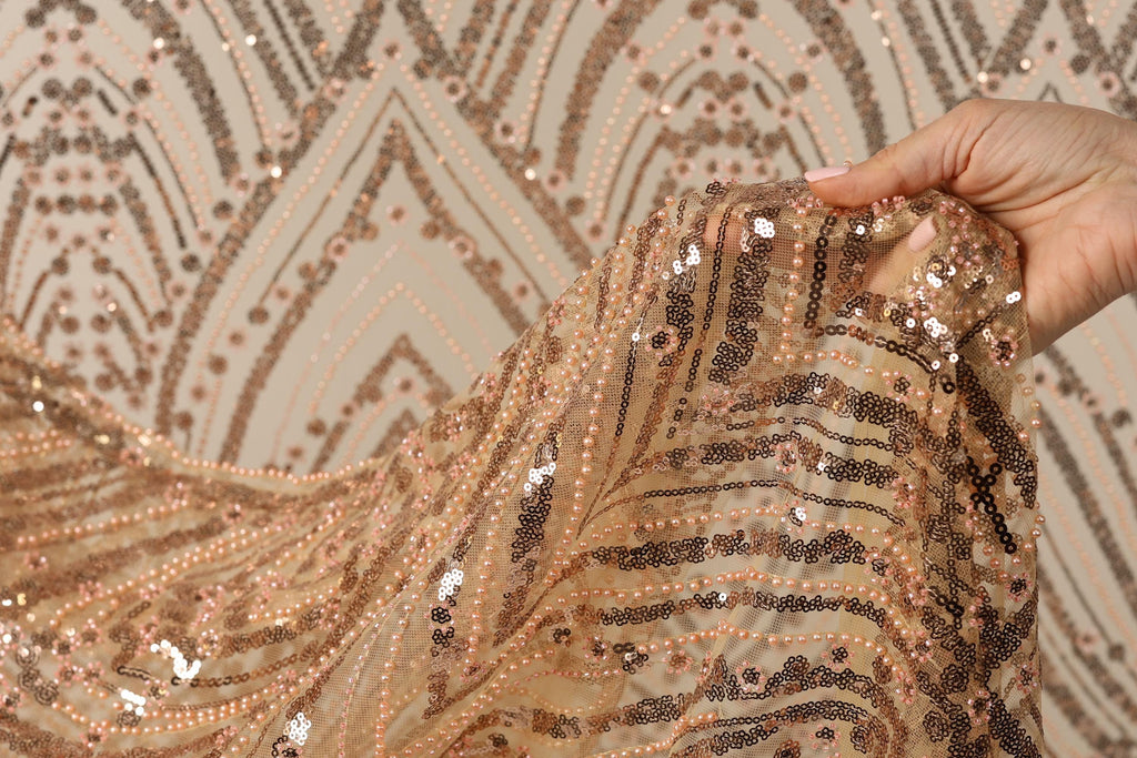 A hand is holding a piece of fabric with a gold and black sequined pattern, against a background with a repeating pattern of the same design.