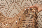 A hand is holding a piece of fabric with a gold and black sequined pattern, against a background with a repeating pattern of the same design.