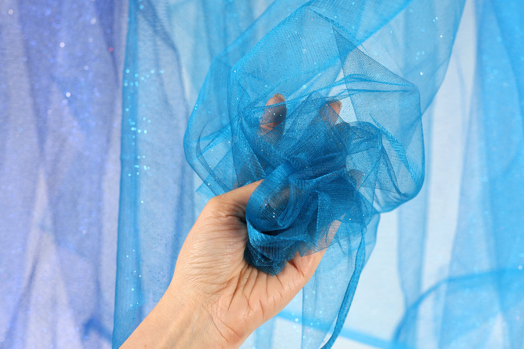 A hand is holding a piece of blue, shiny, and translucent fabric against a blue background.