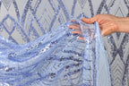 A hand is holding a piece of blue sequin fabric, with the fabric's intricate pattern visible.