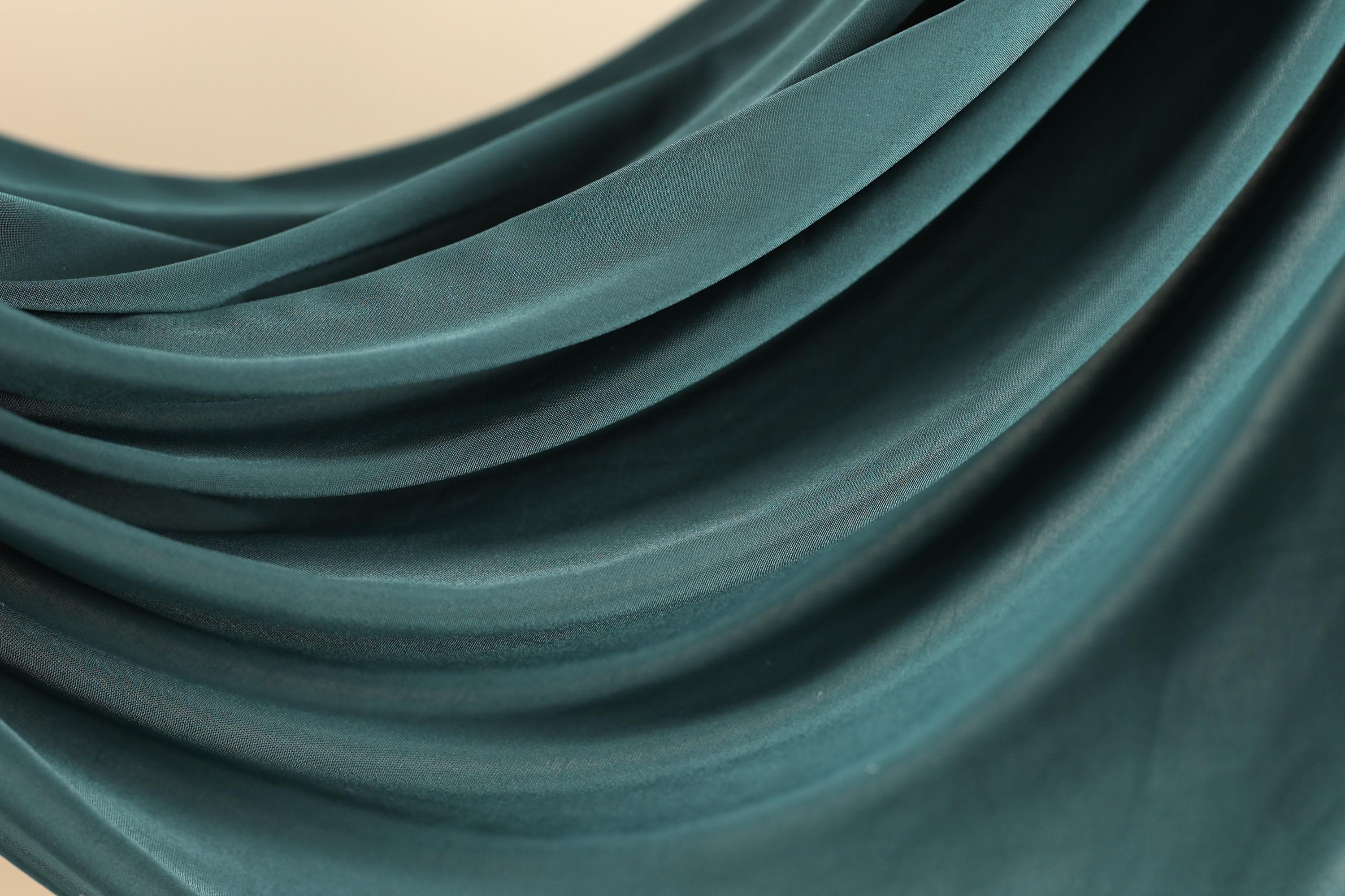 A close-up view of a textured fabric, likely a piece of clothing or textile, with a dark green or teal color. The fabric appears to be made of a soft, flowing material, possibly silk or chiffon, and has a slightly wrinkled appearance.