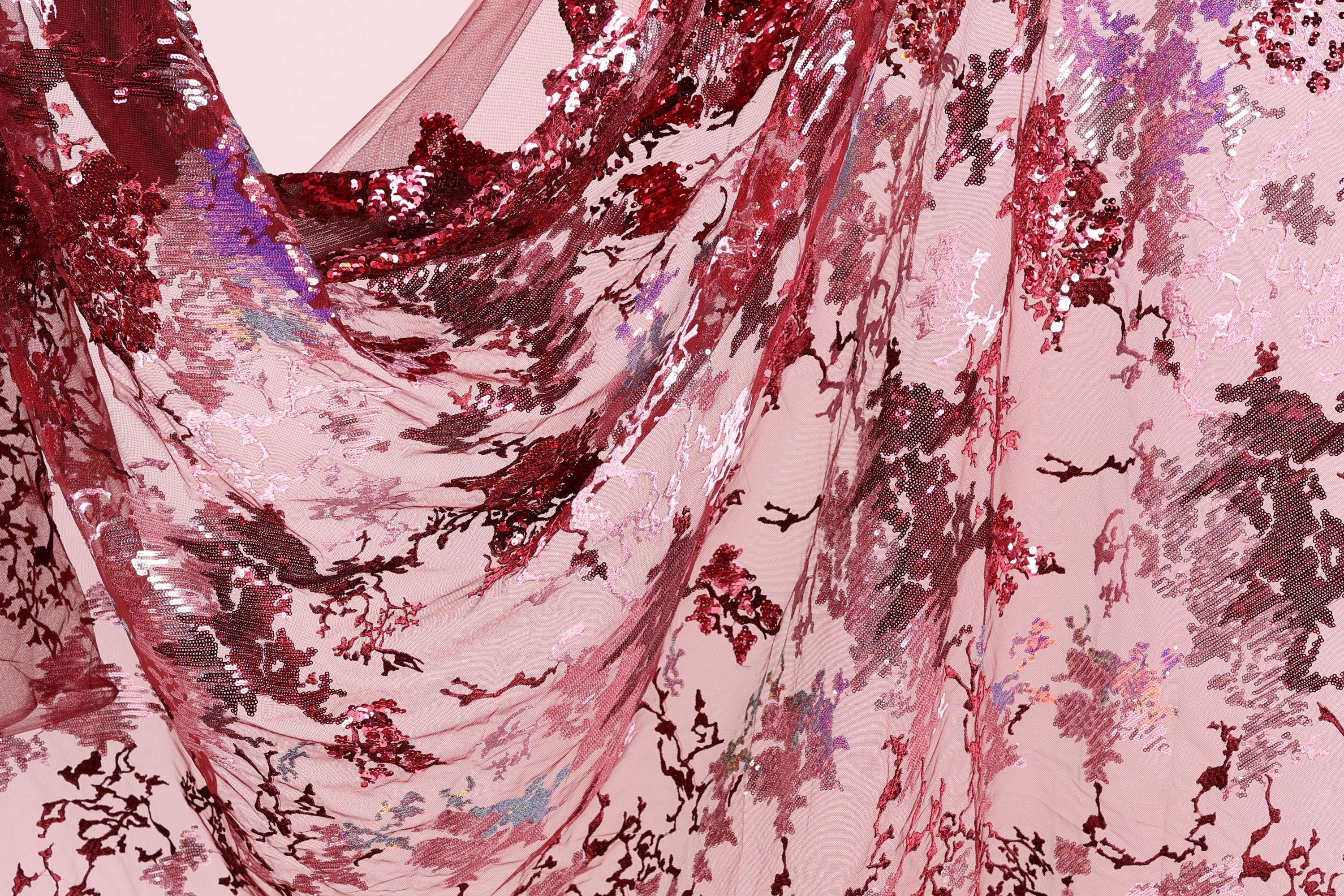 a close-up view of a fabric with a floral pattern in shades of red and pink. The fabric appears to be made of a sheer material, with a delicate and intricate design. The pattern consists of various shades of red and pink flowers and leaves, creating a visually striking and vibrant appearance.