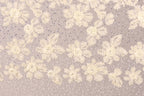 a close-up view of a fabric with a floral pattern. The fabric appears to be made of a light-colored material, possibly lace or a similar type of material. The pattern consists of numerous small white flowers or leaves scattered across the fabric, creating a visually appealing and intricate design. The image does not contain any text or other discernible objects, and the focus is solely on the floral pattern on the fabric.