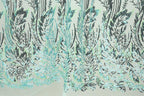 a close-up view of a fabric with a repeating pattern of green and black sequin designs. The pattern appears to be a floral or leaf-like design, with the sequins arranged in a way that creates a sense of depth and texture. The fabric has a light green background, which contrasts with the dark green and black sequin pattern. The image does not contain any text or other discernible objects, and the focus is solely on the intricate and detailed design of the fabric.