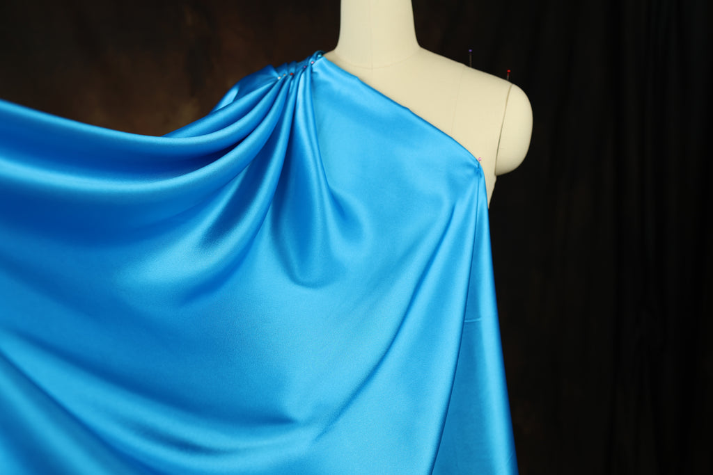 A mannequin wearing a blue satin garment with a draped, flowing fabric draped over its shoulder.