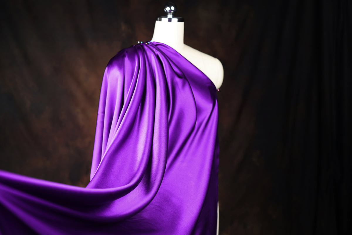 A mannequin wearing a purple satin one-shoulder dress against a dark background.