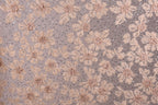 a close-up view of a fabric with a floral pattern. The fabric appears to be a light pink color, with a repeating pattern of small flowers or leaves. The pattern is intricate and detailed, creating a visually appealing texture. The image does not contain any text or other discernible objects, and the focus is solely on the floral pattern on the fabric.
