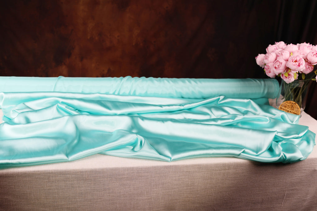 a roll of turquoise satin fabric and a vase of pink roses on a table.