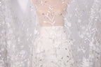 A white dress with a sheer, lace-like fabric draped over it, featuring delicate floral patterns.