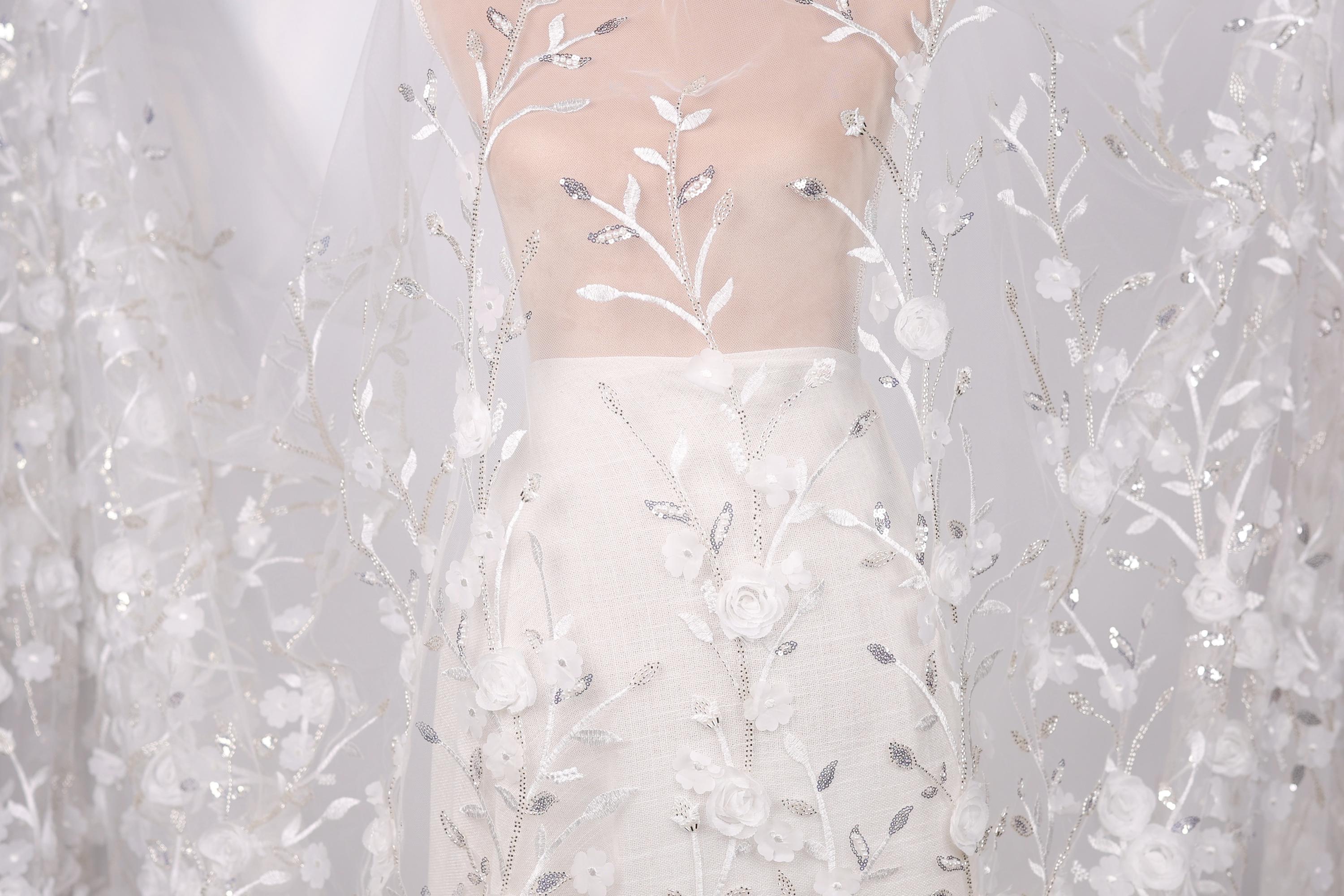 A white dress with a sheer, lace-like fabric draped over it, featuring delicate floral patterns.