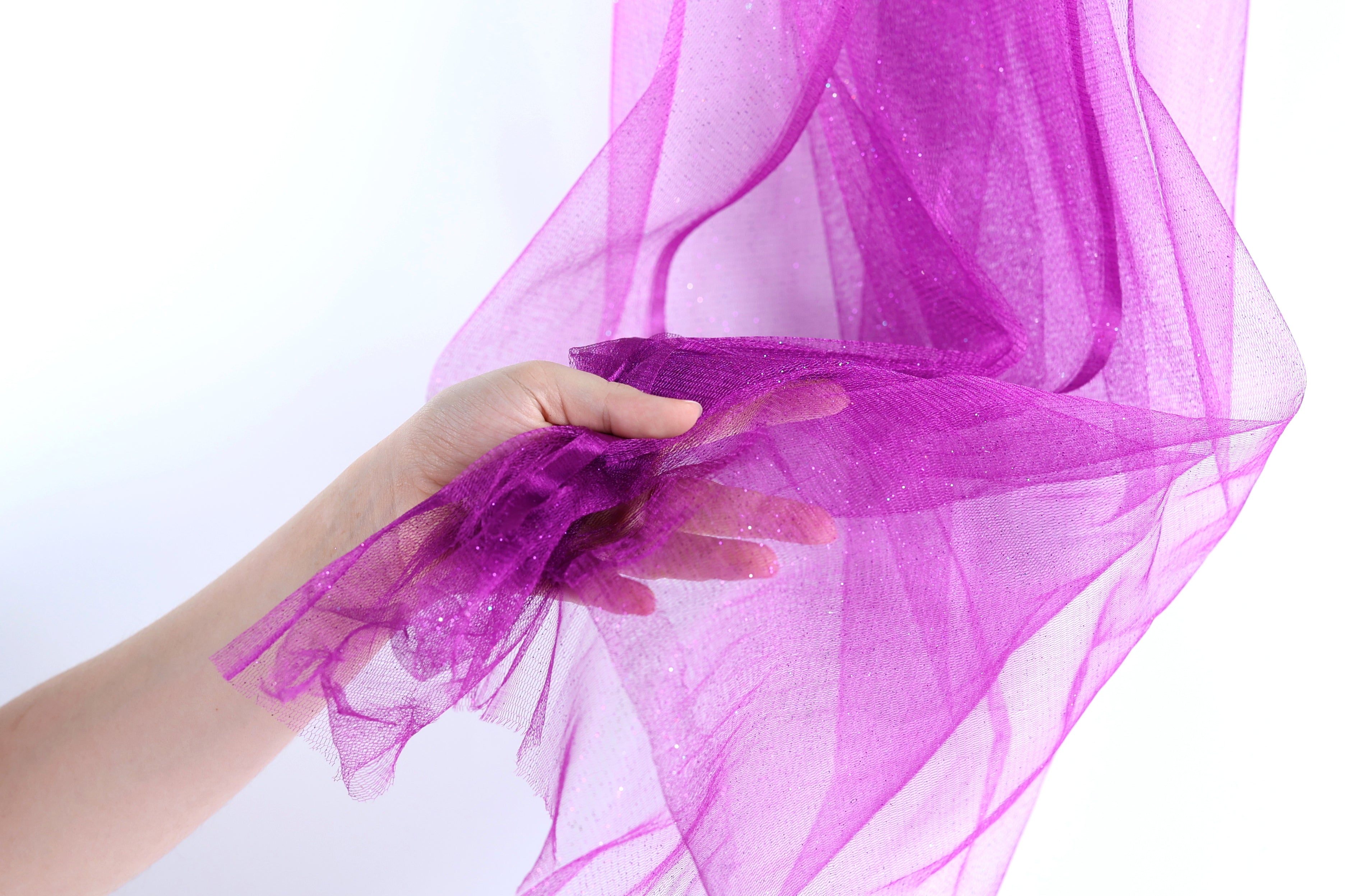 A hand is holding a piece of purple tulle fabric, which appears to be made of a shiny, glittery material.