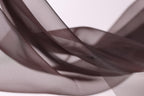 The image is a close-up of a brown fabric, possibly silk or chiffon, with a soft, flowing texture. The fabric appears to be in a crumpled or wrinkled state, giving it a sense of movement and softness. The background is a plain white color, which contrasts with the brown fabric and makes it stand out. The image does not contain any discernible text or countable objects, and the focus is solely on the fabric and its texture.