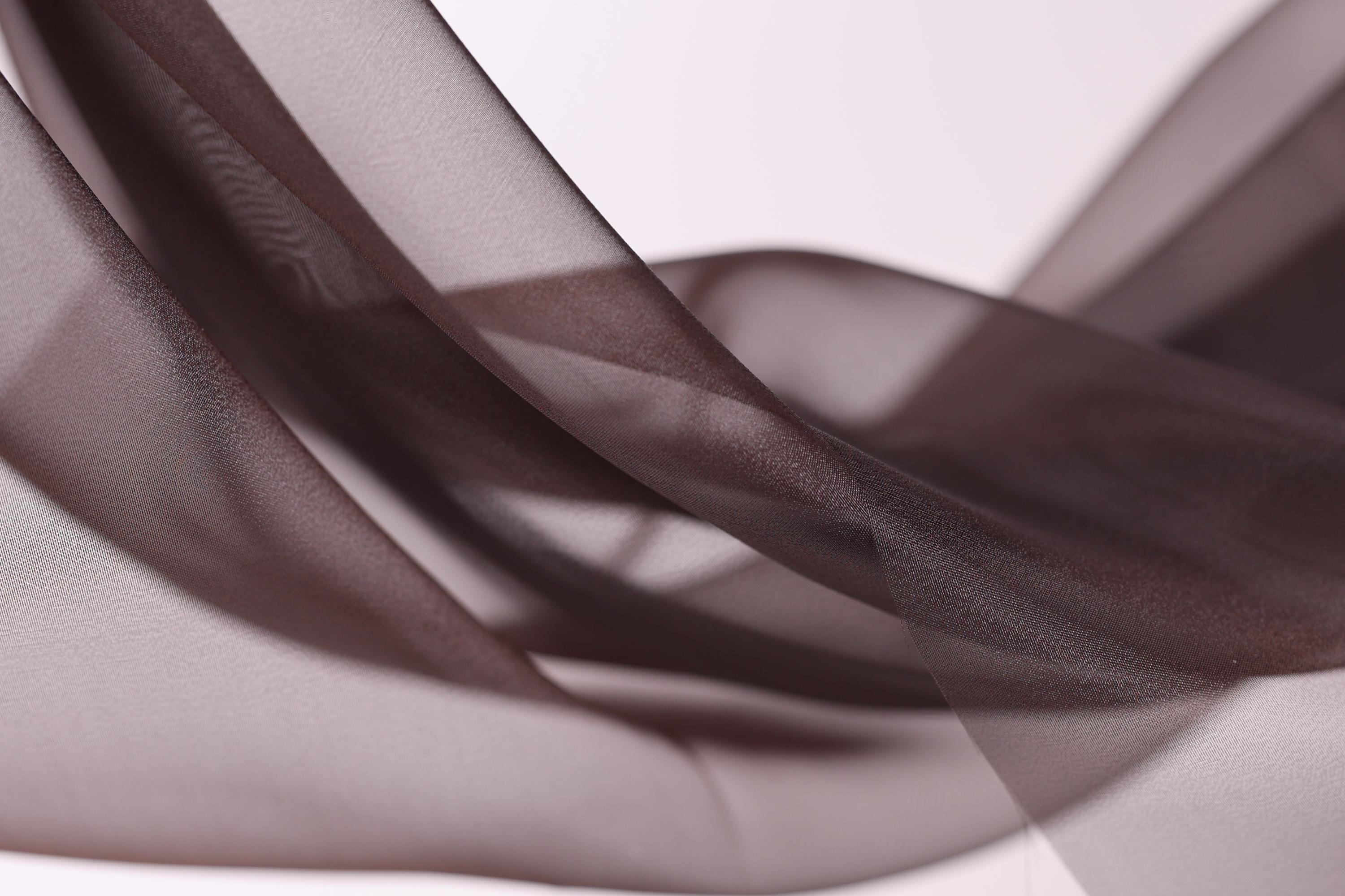 The image is a close-up of a brown fabric, possibly silk or chiffon, with a soft, flowing texture. The fabric appears to be in a crumpled or wrinkled state, giving it a sense of movement and softness. The background is a plain white color, which contrasts with the brown fabric and makes it stand out. The image does not contain any discernible text or countable objects, and the focus is solely on the fabric and its texture.