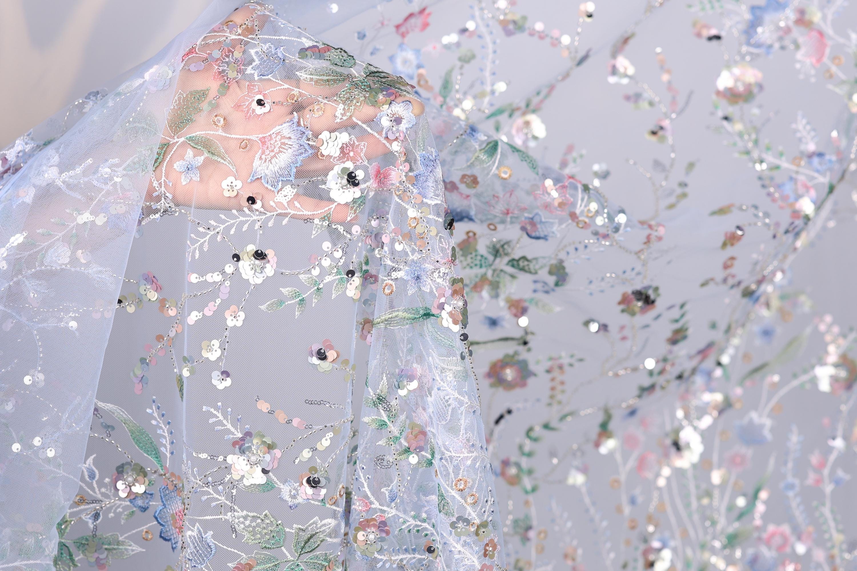 a close-up view of a delicate, intricately embroidered fabric with a floral pattern. The fabric appears to be made of sheer material, allowing the colorful flowers and leaves to be clearly visible. The background is a light blue color, which complements the vibrant colors of the embroidered design. The image does not contain any text or other discernible objects, and the focus is solely on the detailed embroidered pattern.