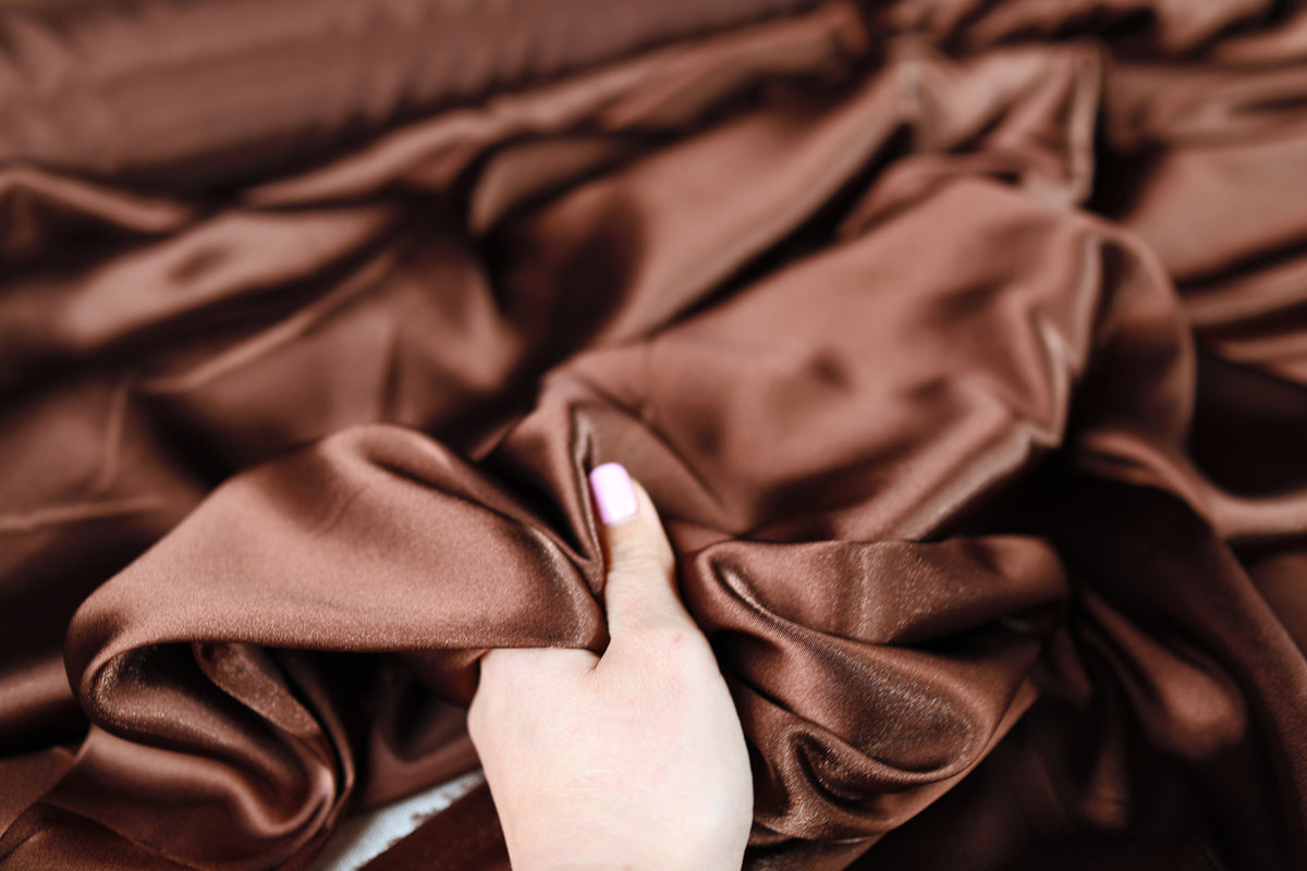 A hand is holding a piece of brown satin fabric, which appears to be a close-up view of the material.