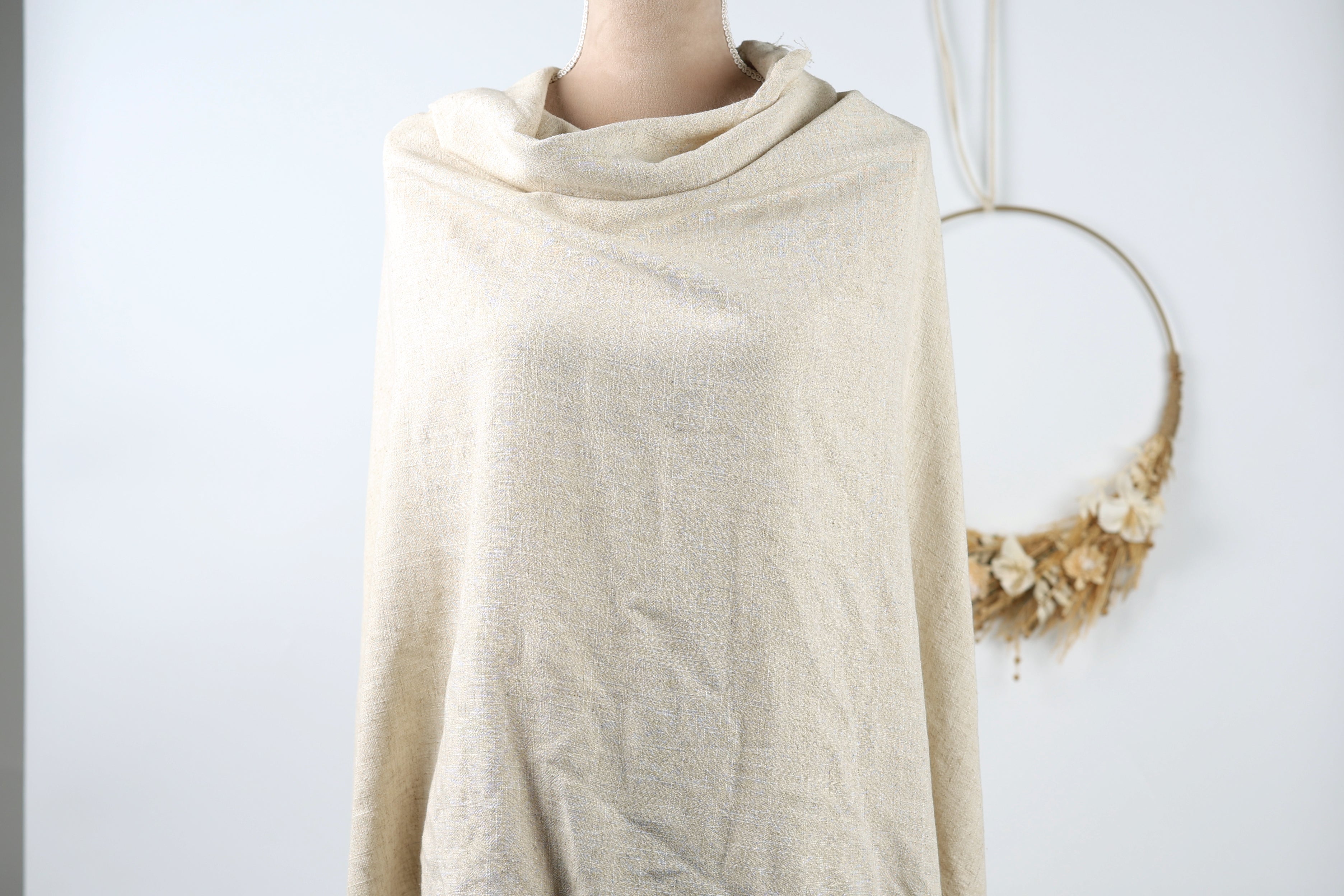 A mannequin wearing a beige shawl stands against a white background, with a wreath hanging on the wall behind it.