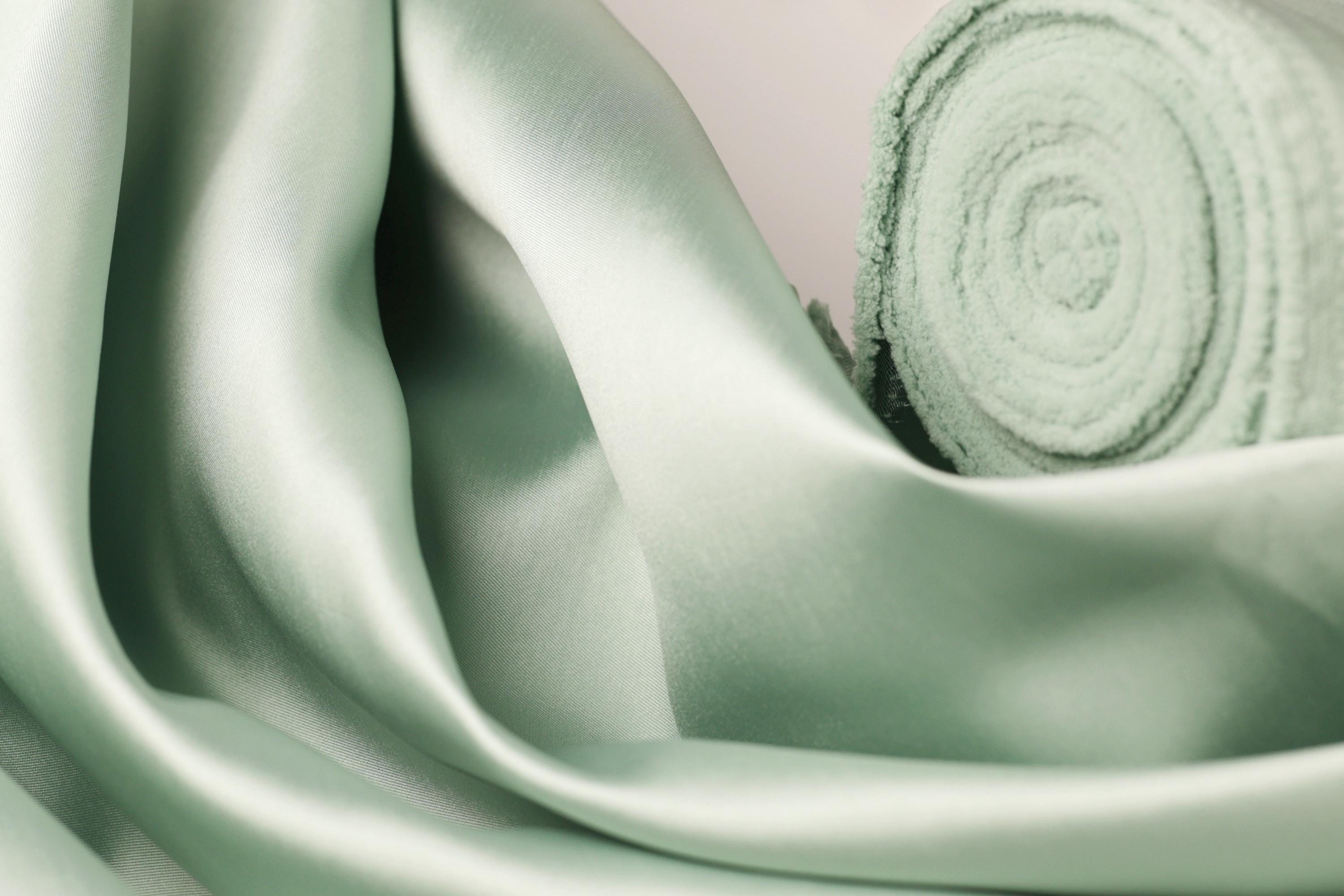 A close-up view of a soft, mint green satin fabric, with a rolled-up piece of the fabric visible in the background.