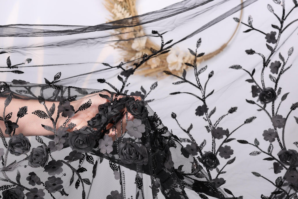 A hand is holding a piece of black lace with a floral pattern, partially obscuring a background of a plant or flower arrangement.