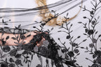 A hand is holding a piece of black lace with a floral pattern, partially obscuring a background of a plant or flower arrangement.