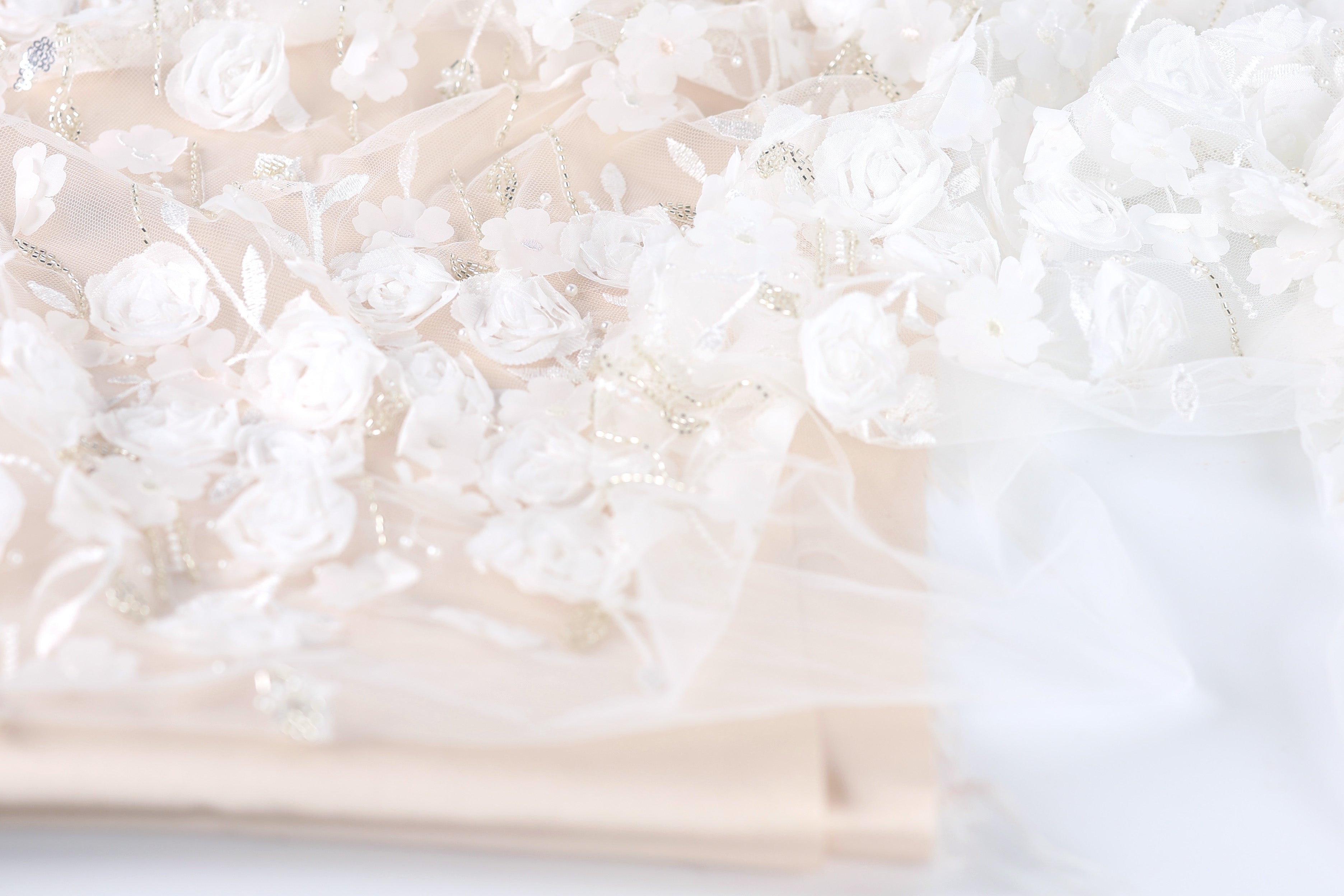 The image is a close-up of a delicate, white lace fabric with intricate floral embroidery. The fabric appears to be a light pink color, and the flowers are arranged in a way that creates a sense of depth and texture. The image does not contain any discernible objects or actions, and the focus is solely on the lace fabric and its floral design.