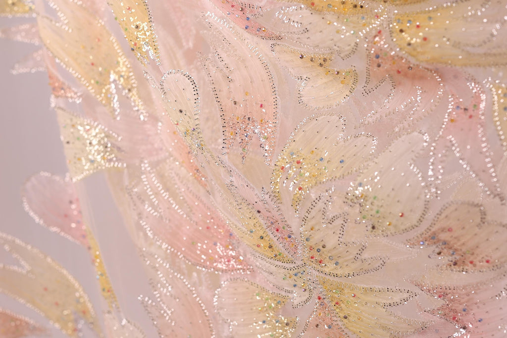 The image is a close-up of a fabric with a floral pattern, featuring a mix of pink, yellow, and white colors. The fabric appears to be made of a shiny, glittery material, adding a sparkly effect to the overall appearance.