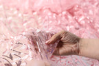 A hand is holding up a piece of pink fabric with sequin details, against a pink background.