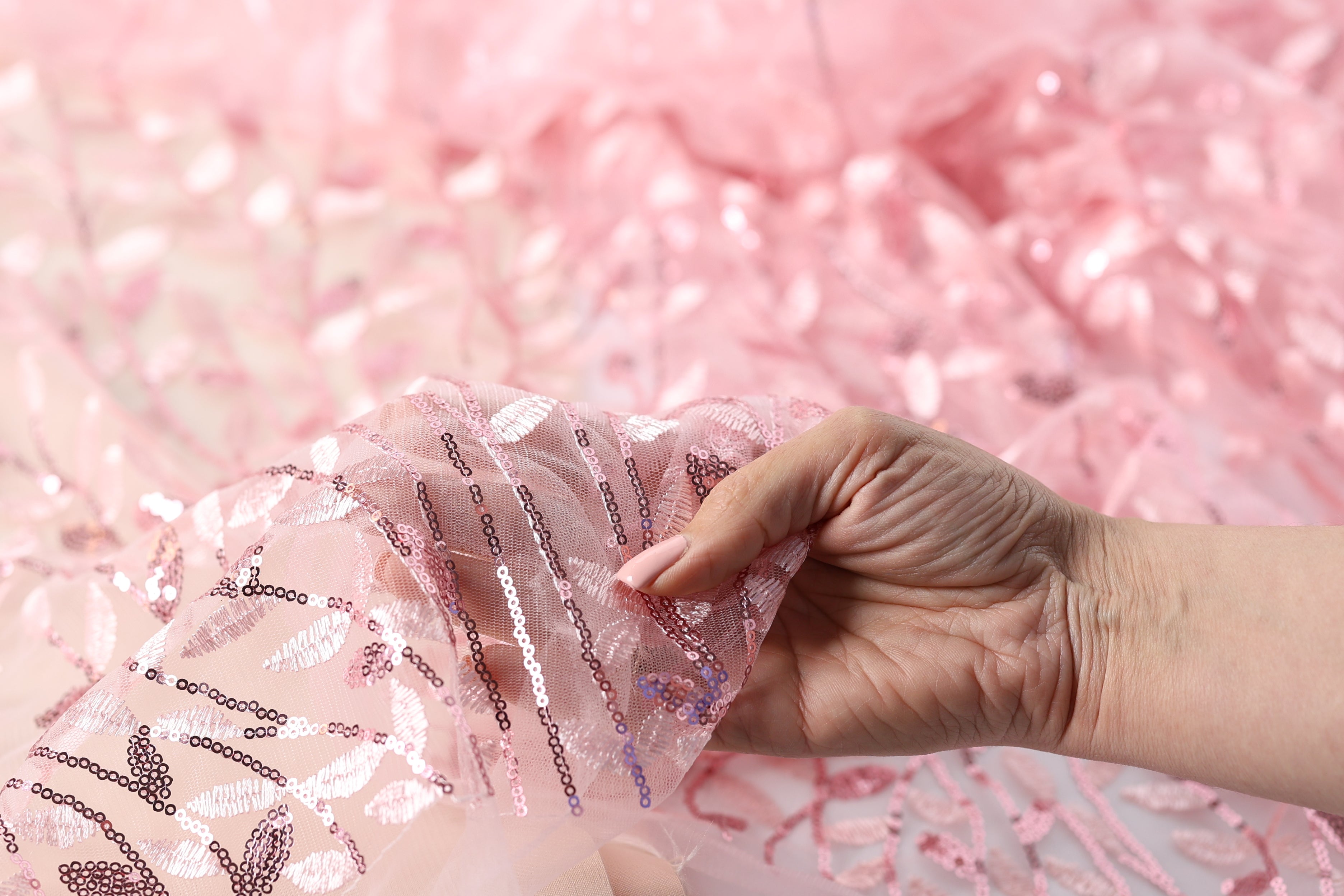 A hand is holding up a piece of pink fabric with sequin details, against a pink background.