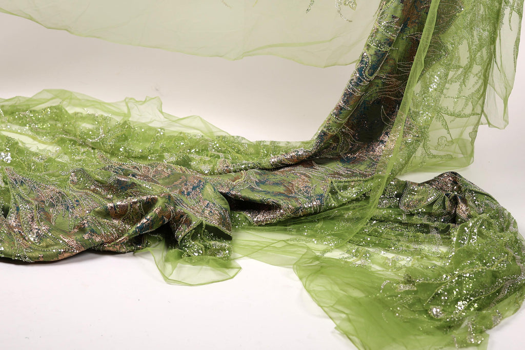 a close-up view of a green, sheer fabric with a patterned design. The fabric appears to be made of a shiny, metallic material and is draped over a surface, creating a visually striking contrast between the green fabric and the white background.