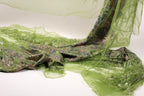 a close-up view of a green, sheer fabric with a patterned design. The fabric appears to be made of a shiny, metallic material and is draped over a surface, creating a visually striking contrast between the green fabric and the white background.
