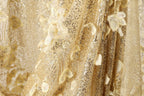 The image is a close-up of a fabric with a gold and white color scheme, featuring a floral pattern and sequin details.