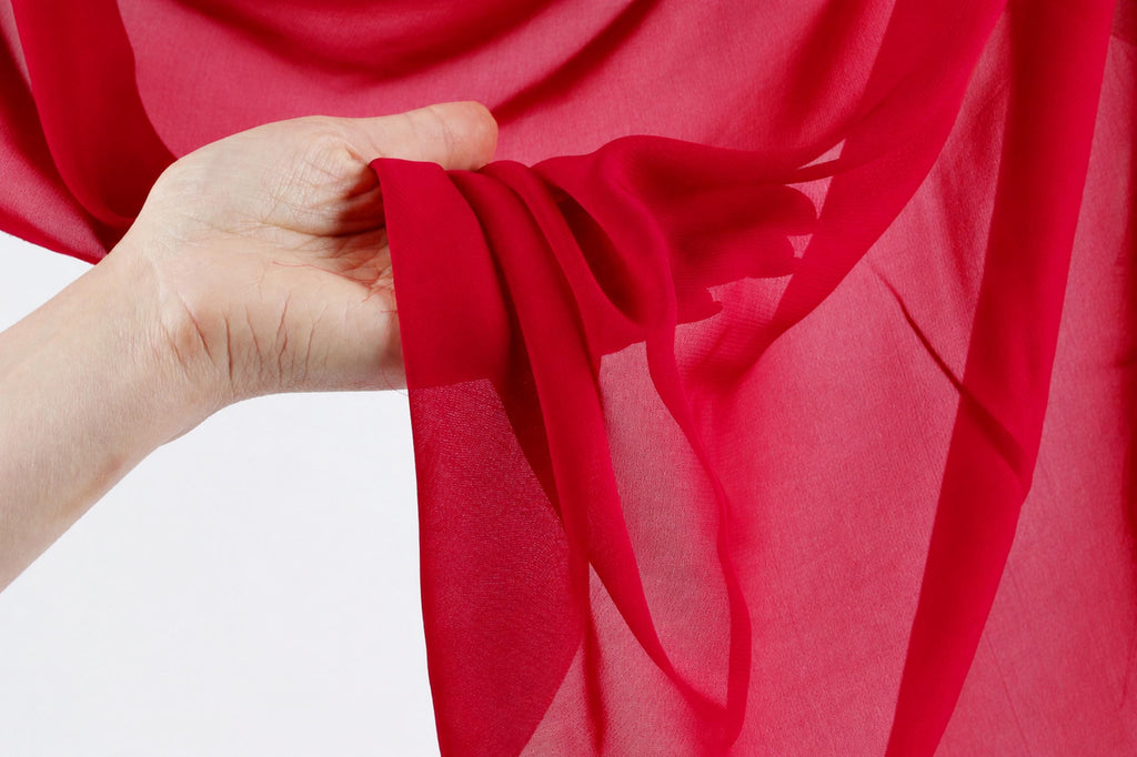 A hand is holding a red fabric, possibly silk or chiffon, against a white background.