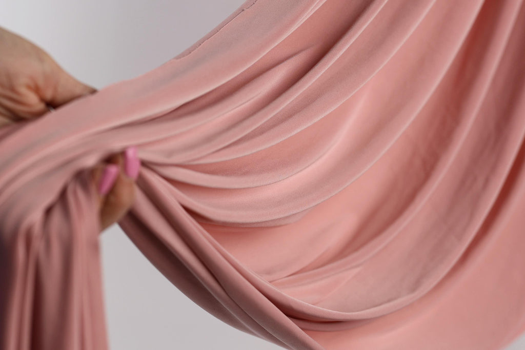 A hand is holding a pink fabric, which appears to be a sheer or sheer-like material. The fabric is being held up against a white background, creating a visually striking contrast.