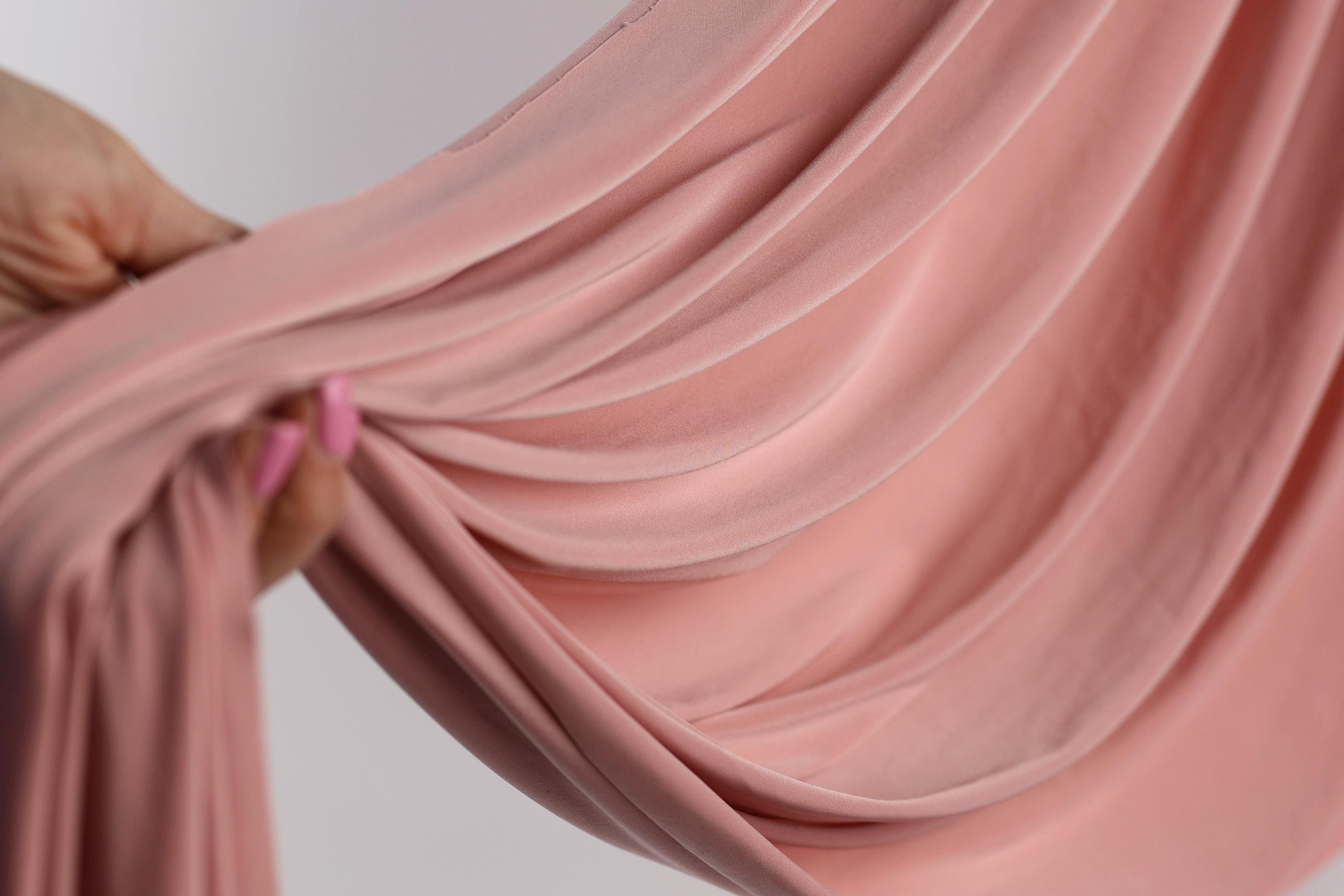 A hand is holding a pink fabric, which appears to be a sheer or sheer-like material. The fabric is being held up against a white background, creating a visually striking contrast.
